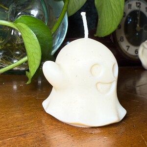 May include: A cream-coloured ghost-shaped candle with a smiling face and a small, curved arm. The candle has a white wick and sits on a wooden surface. Green leaves and a clock are visible in the background.