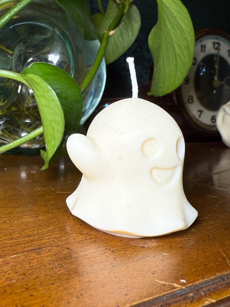 May include: A cream-coloured ghost-shaped candle with a smiling face and a small, curved arm. The candle has a white wick and sits on a wooden surface. Green leaves and a clock are visible in the background.
