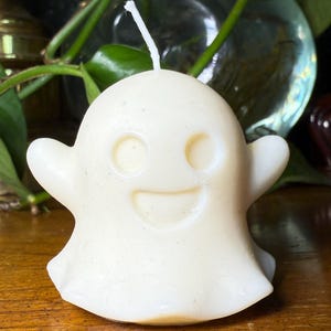 May include: A cream-coloured ghost-shaped candle with a smiling face. The candle has a wick at the top and small arms on each side. The candle is on a wooden surface.