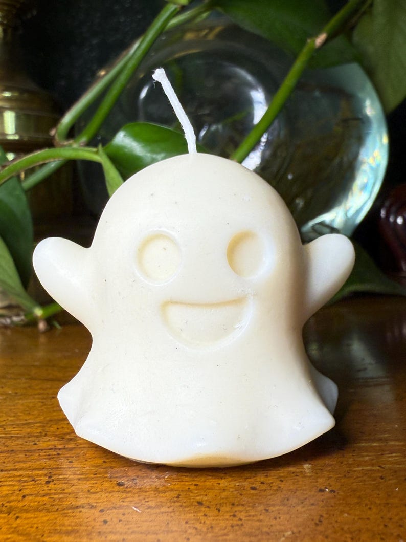 May include: A cream-coloured ghost-shaped candle with a smiling face. The candle has a wick at the top and small arms on each side. The candle is on a wooden surface.