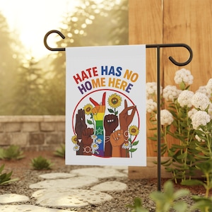 May include: White garden flag with the text "HATE HAS NO HOME HERE". The flag features a rainbow hand gesture, sunflowers, and diverse hands in a circle. The flag is displayed outdoors.