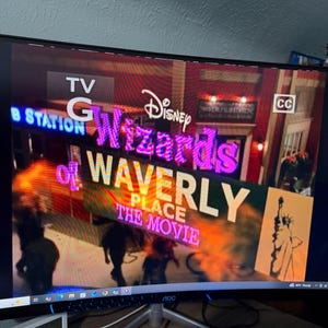 以下が含まれることがあります： ディズニーの「ウェイバリー通りのウィザード: ザ・ムービー」のシーンを表示するコンピューターモニター。タイトルは大きくカラフルな文字で表示されています。画面には、テレビのレーティング、クローズドキャプションのアイコン、ぼやけた人々の画像も表示されています。