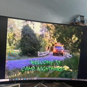 以下が含まれることがあります： コンピューターモニターには、「Camp Nightmare」のシーンが表示されています。画像には、木々に囲まれた車へと続く道が写っています。画面のテキストは、緑と紫で「Welcome to Camp Nightmare」と表示されています。