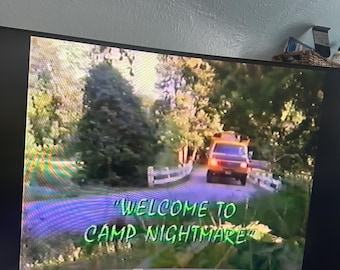 Könnte beinhalten: Ein Computermonitor zeigt eine Szene aus "Camp Nightmare". Das Bild zeigt eine Straße, die zu einem Fahrzeug führt, umgeben von Bäumen. Der Text auf dem Bildschirm lautet "Welcome to Camp Nightmare" in Grün und Lila.