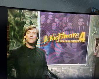 Może przedstawiać: Ekran telewizora wyświetla scenę z filmu "Koszmar z ulicy Wiązów 4". Mężczyzna w ciemnej kurtce stoi przed plakatem z tytułem filmu. Plakat ma fioletowo-czerwony schemat kolorów.
