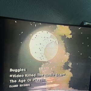 May include: A screen displays the album cover for Buggles' "Video Killed the Radio Star" from "The Age of Plastic" album, released by Island Records. The image features a large, bright circle with a star-filled background.