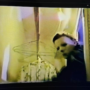 Puede incluir: Imagen borrosa de una persona con una máscara blanca, que recuerda a un personaje de película de terror. La persona está cerca de una puerta, con un fondo borroso. La escena evoca suspense y misterio.