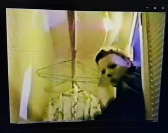 Könnte beinhalten: Ein unscharfes Bild, das eine Person mit einer weißen Maske zeigt, die an eine Horrorfilmfigur erinnert. Die Person befindet sich in der Nähe eines Türrahmens, mit einem verschwommenen Hintergrund. Die Szene erzeugt Spannung und Geheimnis.