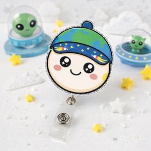 Puede incluir: Un portatarjetas redondo de estilo caricatura con una cara sonriente con un sombrero azul y verde con temática de la Tierra. El portatarjetas tiene un borde plateado brillante y un clip de plástico transparente. Pequeñas decoraciones con forma de estrella y extraterrestre están dispersas.