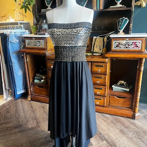 May include: A strapless black dress on a mannequin. The bodice is embellished with horizontal bands of silver sequins and gold beading. The skirt is a flowing, floor-length design. The dress is displayed in a vintage shop setting.