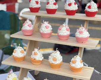 May include: A wooden tiered display holds several cupcake-shaped candles. The candles have white frosting and are in red, pink, orange, and yellow cupcake liners. Some are decorated with heart-shaped sprinkles.