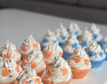 May include: A collection of cupcake-shaped candles with white frosting, orange heart decorations, and sprinkles. The candles are in orange and blue cupcake cups, arranged on a white surface.