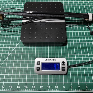 以下が含まれることがあります： 黒い電子機器とコイル状のコード、およびベージュの円筒形部品が黒いデジタルスケールの上に置かれています。スケールのディスプレイには4.7オンスの重量が表示されています。この機器は、長く質感のある黒いアームと金属製のコネクタを備えています。