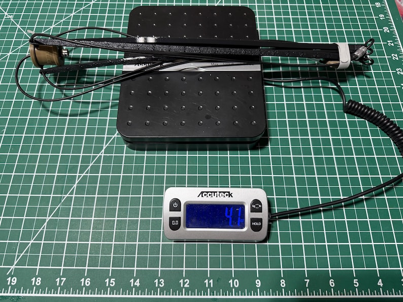 以下が含まれることがあります： 黒い電子機器とコイル状のコード、およびベージュの円筒形部品が黒いデジタルスケールの上に置かれています。スケールのディスプレイには4.7オンスの重量が表示されています。この機器は、長く質感のある黒いアームと金属製のコネクタを備えています。