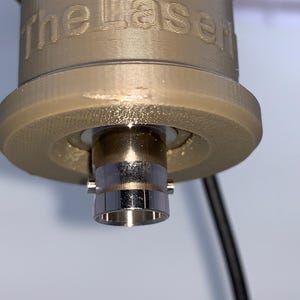 以下が含まれることがあります： 側面に「The Laser」と刻印された、ベージュ色の円筒形3Dプリントハウジング。ハウジングの下部からは銀色の金属製BNCコネクタが突き出ており、側面には細い黒色のケーブルが伸びている。
