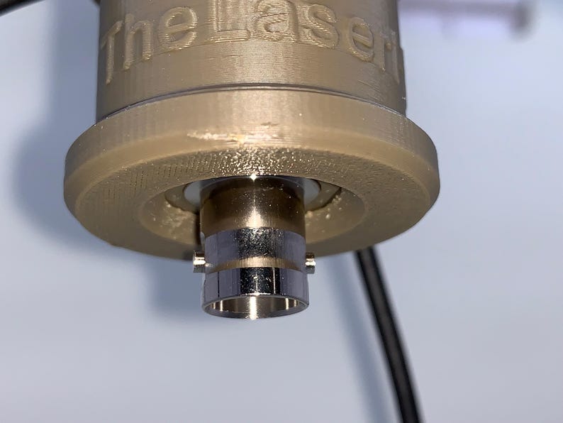 以下が含まれることがあります： 側面に「The Laser」と刻印された、ベージュ色の円筒形3Dプリントハウジング。ハウジングの下部からは銀色の金属製BNCコネクタが突き出ており、側面には細い黒色のケーブルが伸びている。