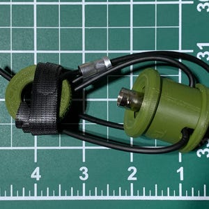 May include: A green and black cable connector with a black strap. The connector has a silver metal tip and a black wire attached to it.