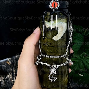 May include: A glass jar with a black lid and a red eye on top. Inside the jar is a preserved snake with a green and white body. The jar is decorated with a silver chain and a skeleton pendant.
