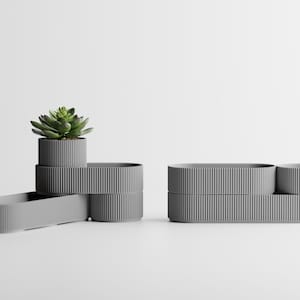 May include: A set of modular gray planters with a vertical ribbed texture. The design consists of multiple interconnected oval and circular segments, with one small green succulent plant placed inside the top circular section of the left unit.