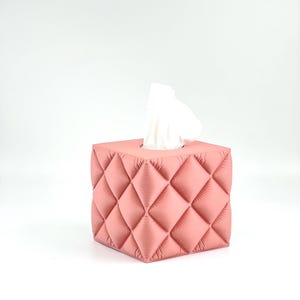May include: A square tissue box cover in a dusty rose pink colour featuring a quilted, diamond-patterned texture with puffed, padded sections. A single white tissue is pulled through the rectangular opening at the top of the box.