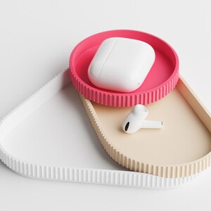 May include: A modern, minimalist tray set in white, beige, and pink. The set includes a white tray, a beige tray, and a pink tray. A white wireless earbud case and a single earbud rest on the trays.