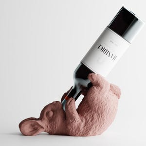 May include: A wine bottle held by a pink bunny-shaped wine bottle holder. The wine bottle has a white label with black text. The bunny is lying on its back, appearing to drink from the bottle. The background is white.