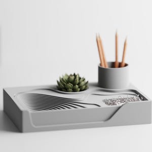 Puede incluir: Un organizador de escritorio gris con un plantador de suculentas incorporado, un portalápices y una bandeja para clips. El organizador contiene varios lápices y una pequeña suculenta verde. El diseño presenta un patrón ondulado.