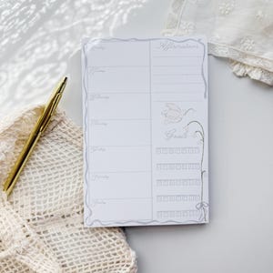 May include: A white weekly planner notepad featuring a gray border, a gold pen, and sections labeled Affirmations and Goals. The planner includes days of the week from Monday to Sunday, a small bird illustration, and rows of boxes with letters M, T, W, T, F, S, S.