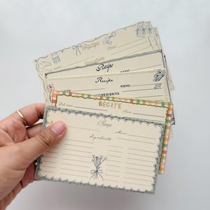 Puede incluir: Un montón de tarjetas de recetas de estilo vintage con bordes decorativos y texto escrito a mano. Las tarjetas presentan secciones para el nombre de la receta, los ingredientes y el memorándum. La paleta de colores incluye crema, azul y acentos pastel, con ilustraciones florales y de lazos.