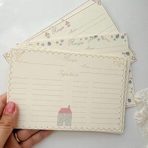 May include: A stack of cream-colored recipe cards with scalloped edges. The top card features a small illustration of a house with a pink roof, the word Recipe, and lines for Dish name, Mimi, and Ingredients. Other cards show floral and dot patterns.