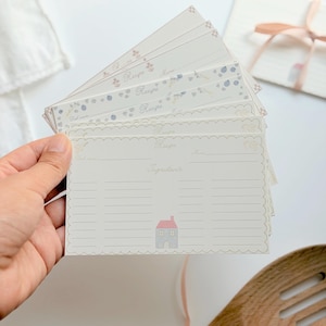 May include: A stack of cream-colored recipe cards with scalloped edges. Each card features the printed text 'Recipe', 'Ingredients', 'Date', and 'Name', along with a small illustration of a house with a pink roof and blue walls.
