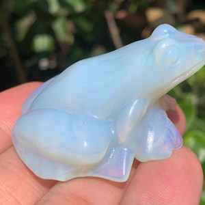 May include: A small, translucent, light blue opalite figurine carved in the shape of a frog. The material has a smooth, polished surface with an iridescent sheen that reflects soft white and pale blue light across its curved, sculpted body.