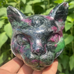 May include: A carved cat head sculpture made of a stone with a mix of colors. The sculpture has a black base color with pink and green accents. The cat's features are well-defined, with visible eyes, nose, and ears. The sculpture is held in a hand.