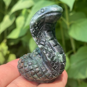 May include: A carved stone snake figurine in shades of green and black. The snake is coiled, with its head raised, and features detailed scales. The background is blurred green foliage.