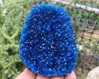 May include: A vibrant blue crystal geode, showcasing a dense cluster of sparkling, pointed crystals. The geode is held in a hand, with a blurred green background.