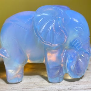May include: A small, light blue glass elephant figurine. The elephant is standing with its trunk raised and its head tilted to the side. The figurine is made of opalescent glass, which gives it a shimmering, iridescent appearance.