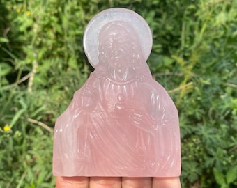 May include: A pink rose quartz carving of Jesus Christ with a halo. The carving is detailed and shows Jesus with his hands clasped in prayer.