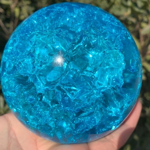 May include: A clear, spherical object with a cracked, textured surface. The sphere is a vibrant shade of blue, with light reflecting off the facets. The object is held in a hand, with a blurred green background.
