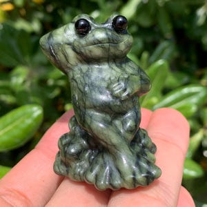Pode incluir: Pequena estatueta de rã verde com olhos pretos. A rã é esculpida em pedra com um padrão mosqueado. A rã está em pé com os braços cruzados. O fundo é folhagem verde desfocada.