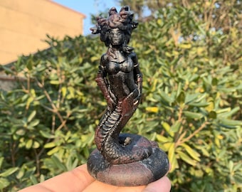 May include: A dark sculpture of Medusa, a figure from Greek mythology. The statue depicts a woman with a snake-like lower body and a head of snakes. The sculpture is mainly black with reddish-brown accents and stands on a circular base.