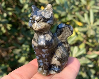 May include: A small, carved stone cat figurine. The cat is gray, black, and blue with a speckled pattern. It is standing upright with its tail curled. The figurine is held in a hand against a blurred green background.