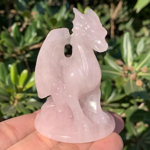 May include: A light pink rose quartz dragon figurine with detailed wings and a textured surface. The dragon is in a seated position, with its wings spread. The figurine is a decorative item, perfect for crystal collectors.