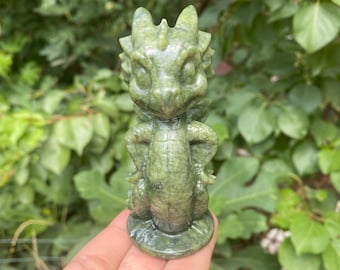 Op de afbeelding: Een groene drakenfiguur met gedetailleerde schubben en een vriendelijke uitdrukking. De draak is gesneden uit een jade-achtige steen en is ongeveer 10 cm hoog. Het beeldje staat tegen een achtergrond van groen gebladerte.