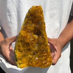 Könnte beinhalten: Eine große, dreieckige Kristallstufe aus durchscheinenden, tief bernsteinfarbenen Kristallen. Das Mineralexemplar weist zahlreiche gezackte, facettierte Spitzen auf, die das Licht einfangen und einen lebendigen, warmen gelben und orangen Glanz erzeugen.