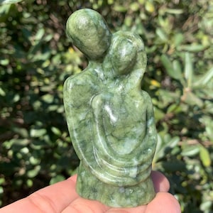 Può includere: Una scultura in pietra verde raffigurante due figure che si abbracciano. La scultura lucida è tenuta in mano, su uno sfondo di fogliame verde. Le figure intrecciate suggeriscono un tema di amore o connessione, con una finitura liscia.