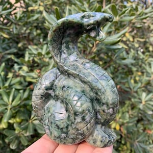 May include: A carved green serpentine stone sculpture of a cobra with its hood raised and mouth open. The snake is coiled, and the stone has a mottled pattern of dark and light green.