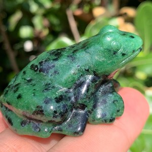 Può includere: Una statuetta di rana scolpita nella pietra con una superficie lucida di colore verde brillante e macchie nere scure sparse. La scultura raffigura una rana in posizione seduta con occhi e zampe ben definiti.
