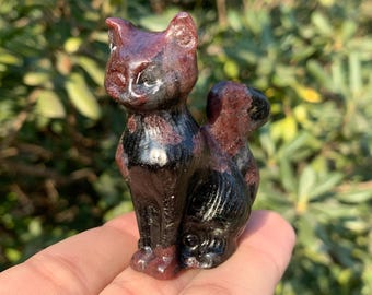 May include: A carved stone cat figurine. The cat is in a sitting position with its tail curled up. The stone is a mix of dark red and black, with a glossy finish. The figurine is held in a hand.