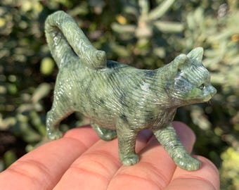 May include: A green and gray stone cat figurine. The cat is in a walking pose with its tail curved over its back. The figurine is made of a stone material with natural variations in color and texture. The cat's features are detailed, including its ears, eyes, and paws.