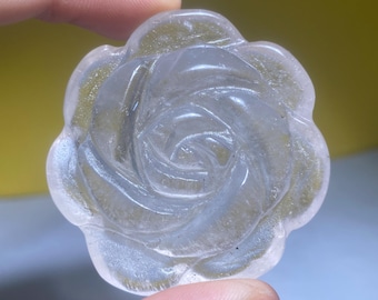 May include: A clear, carved rose-shaped crystal. The crystal is held between two fingers and has a detailed floral design. The petals are translucent, and the overall appearance is smooth and polished. The background is a soft yellow.